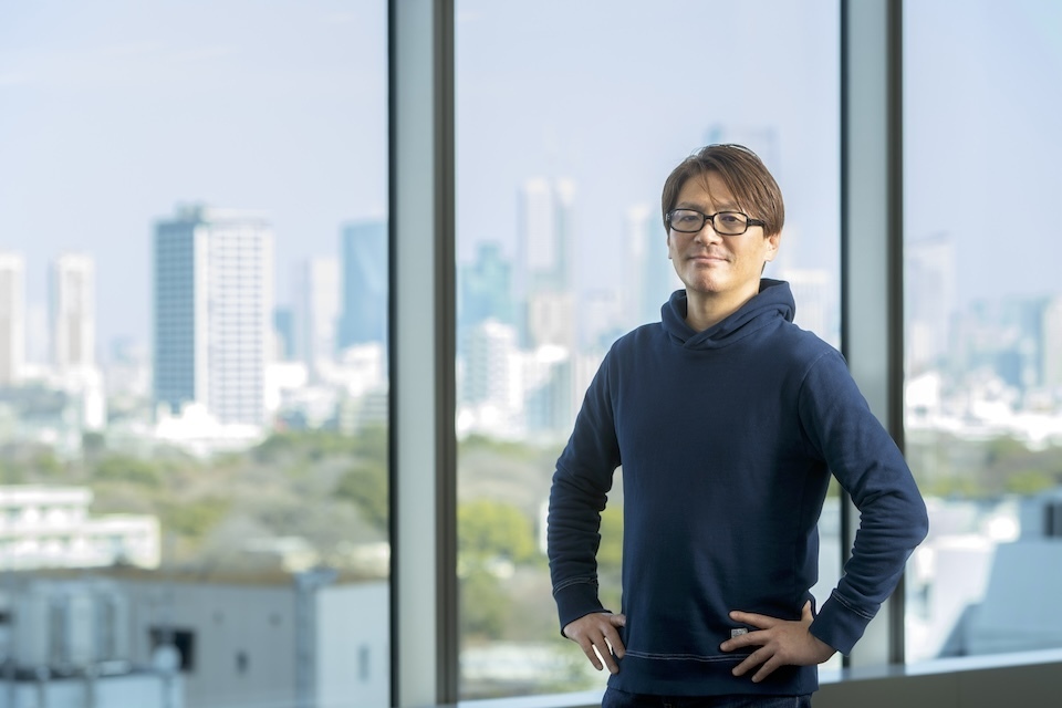 A bespectacled man stands in front of a window, with a bustling city skyline visible behind him.