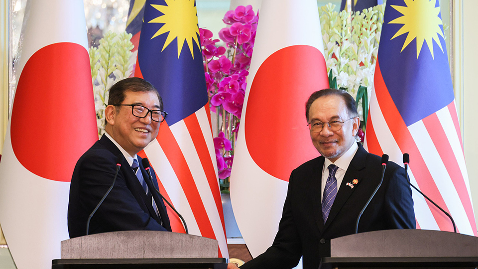 Two men standing behind two podiums, engaged in a handshake, smiling, with Malaysian and Japanese flags displayed prominently in the background.