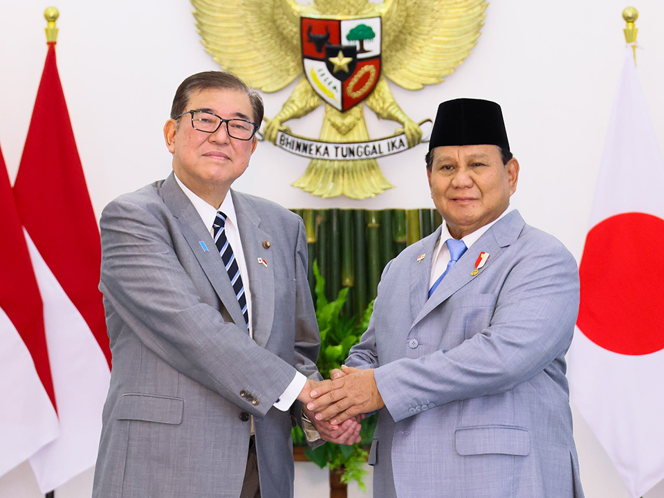 Two men engaged in a handshake, with Indonesia and Japan flags in the background. One is wearing a traditional Indonesian men's hat.