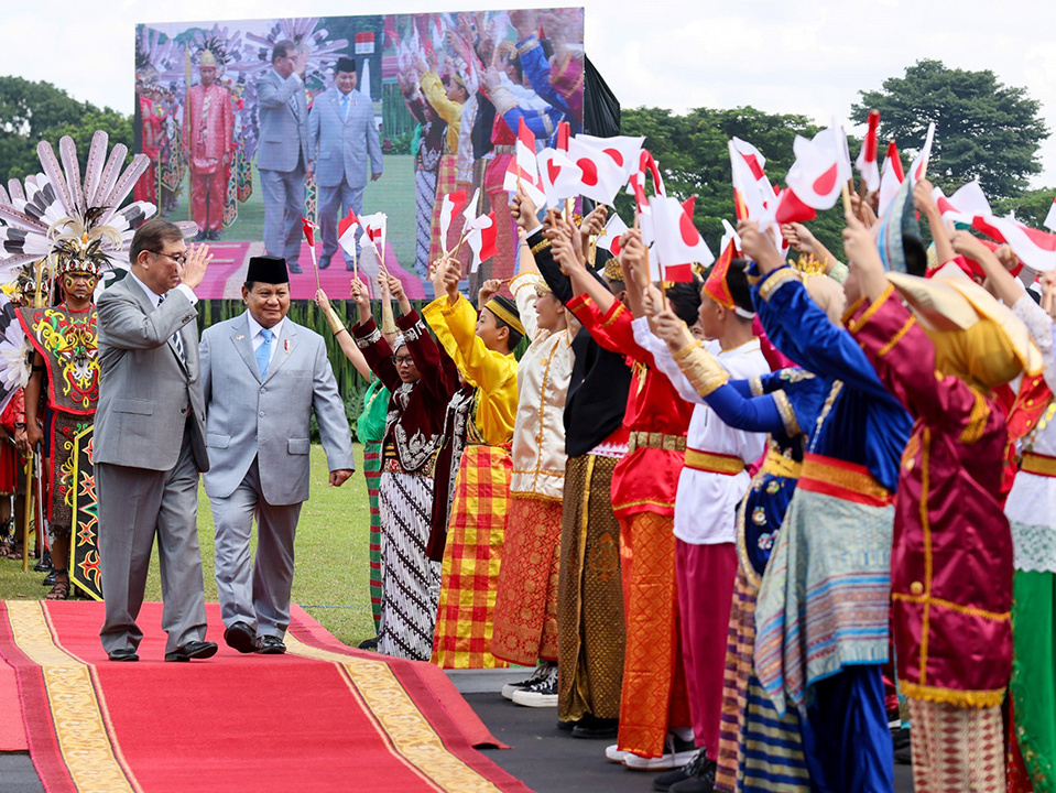 Prime Minister Ishiba waving at audiences in traditional uniforms as part of a welcome ceremony on a red carpet.