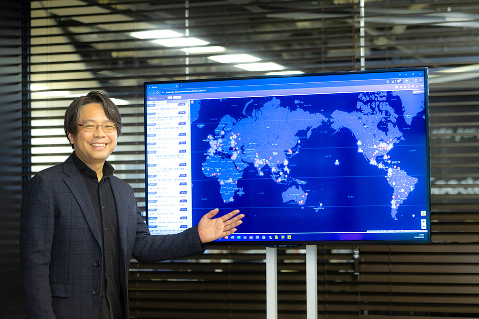 Murakami Kenjiro, CEO of Spectee Inc., stands in front of a large screen displaying Spectee Pro, with a blue world map.