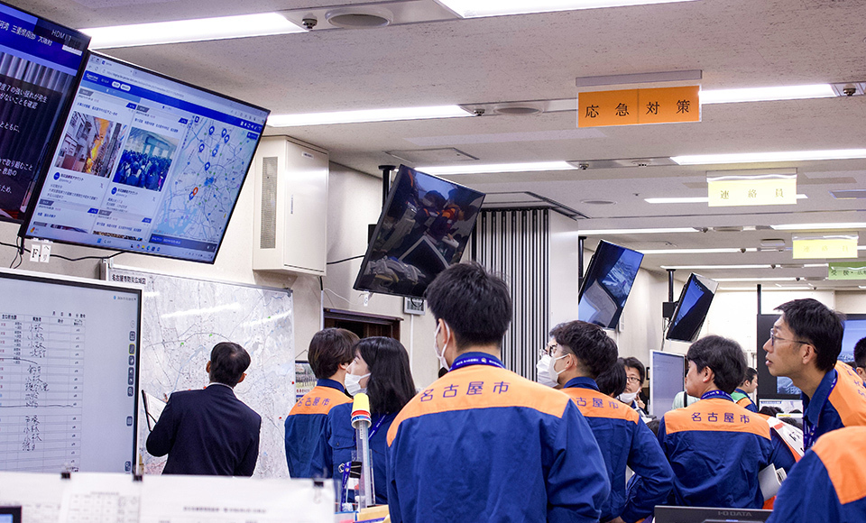 A group of individuals in an office setting, wearing orange and blue uniforms, engaged in a disaster drill with large screens displaying Spectee Pro in Nagoya City, Aichi Prefecture.