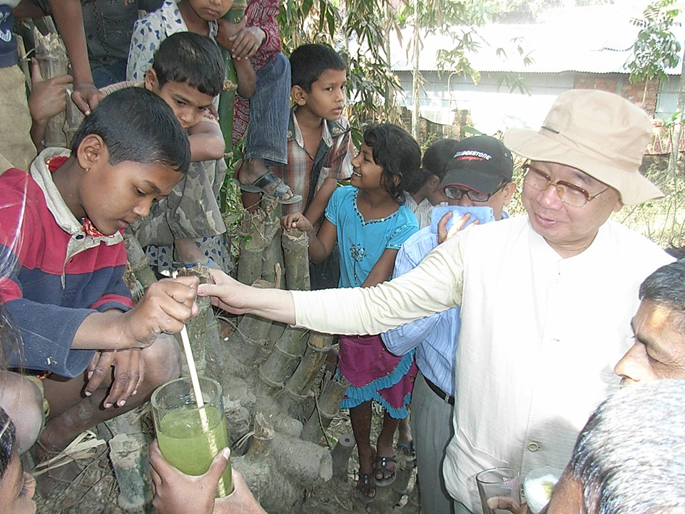 Man educating residents in Bangladesh about water purification as a child stirs green water.