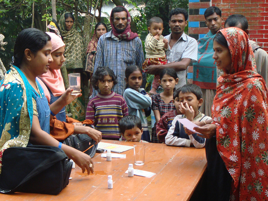 A woman in traditional Bangladeshi clothes stands by a table where two women demonstrate, with a group gathered around them.