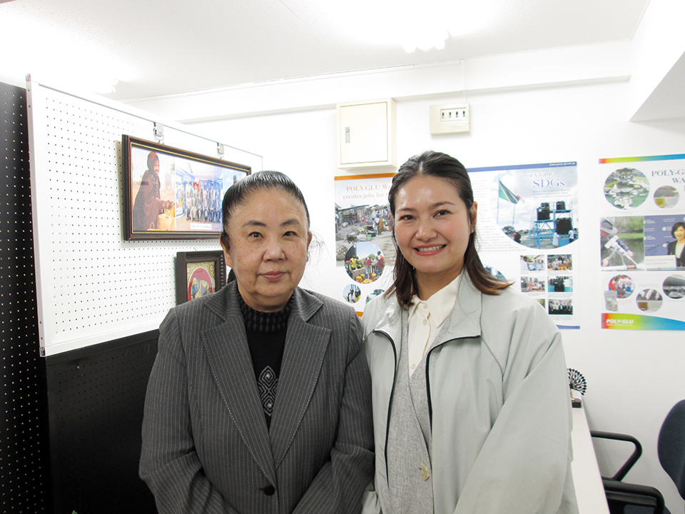 Two women standing in an office, smiling at the camera with posters visible on the wall behind them.